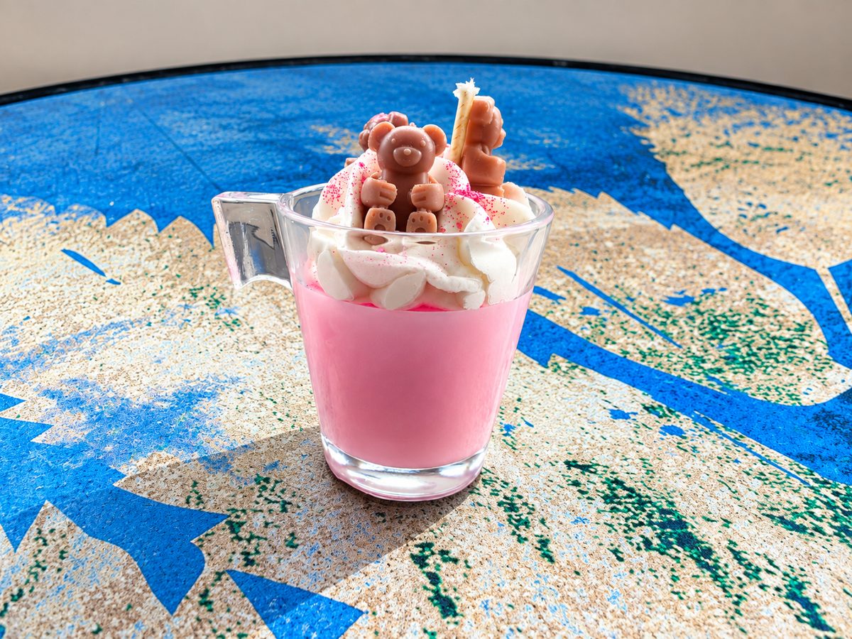 A pink dessert in a clear glass mug topped with whipped cream, marshmallows, and small chocolate teddy bear candies, set on a blue and beige patterned surface.