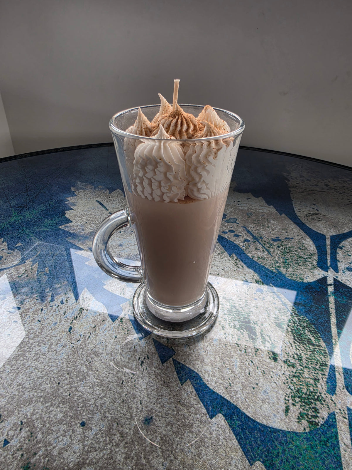 A tall clear glass mug filled with a light brown beverage, topped with piped whipped cream and a small pastry decoration, set on a patterned blue and gray table against a plain gray wall.