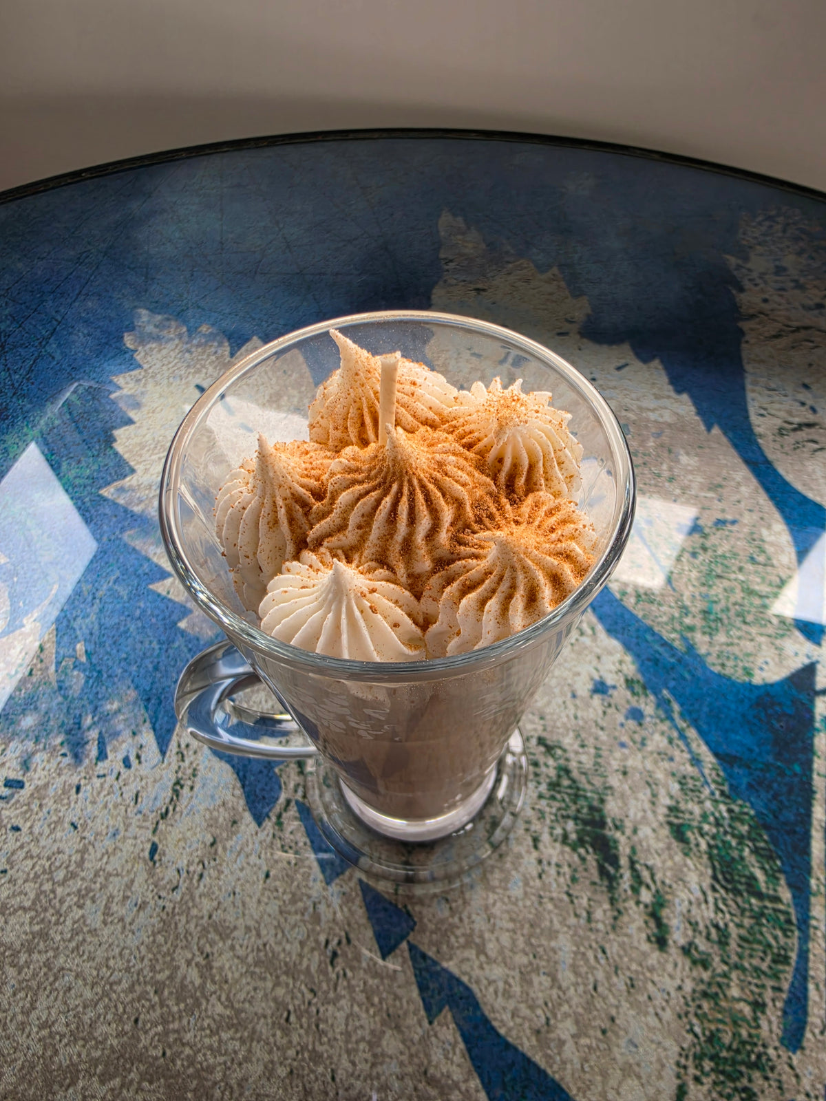 A clear glass mug filled with piped beige meringue-like pastries or whipped cream, set on a reflective blue and gray tabletop.