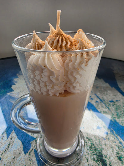 A tall clear glass mug filled with a light brown beverage, topped with piped whipped cream and a sprinkle of cinnamon, set on a blue and white patterned surface.