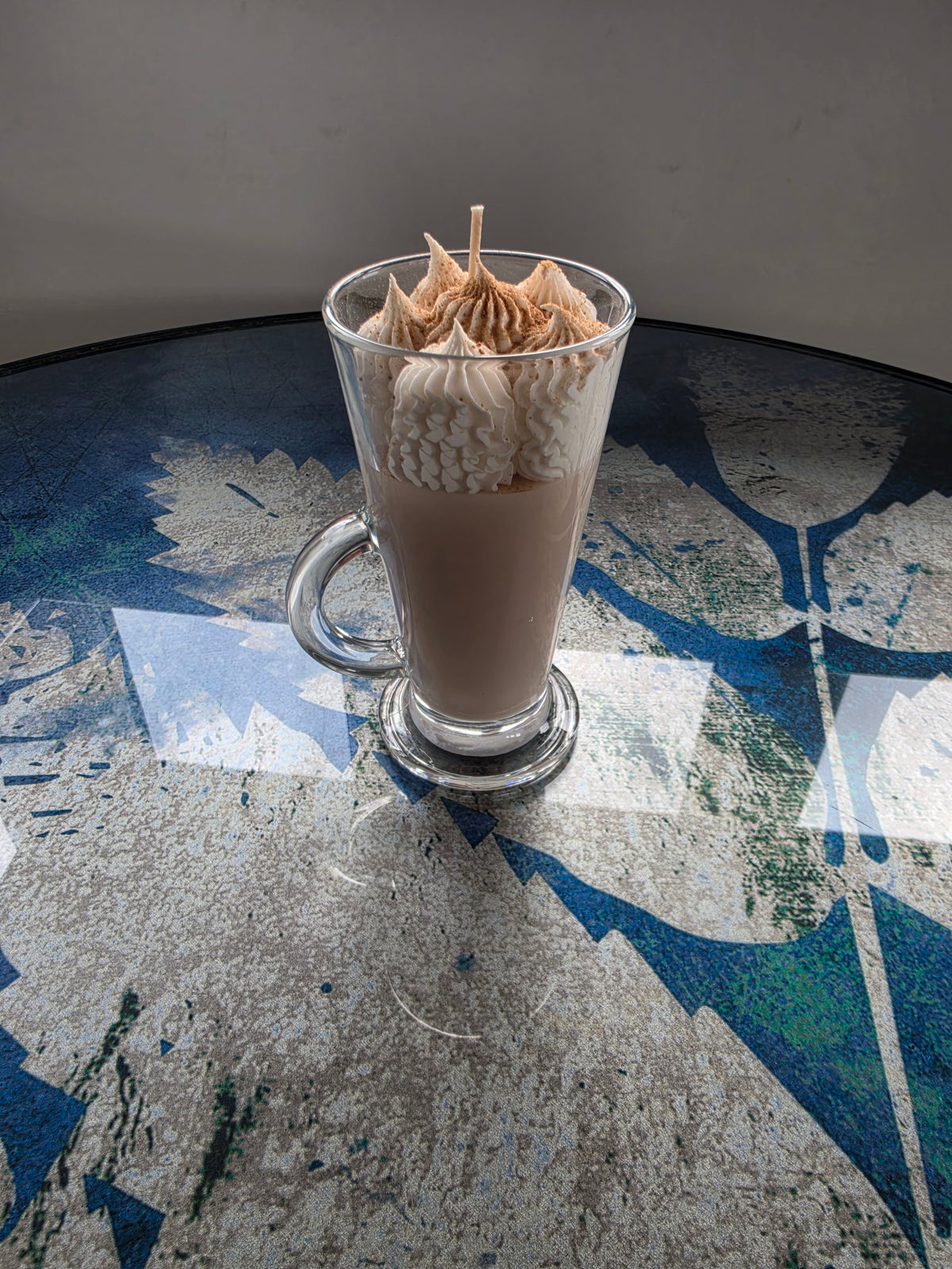 A clear glass handle mug filled with a light brown beverage, topped with piped whipped cream and a small wooden stick, set on a reflective blue and gray patterned tabletop.