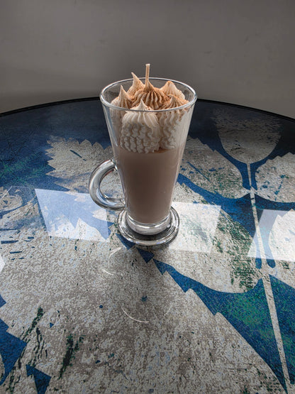 A clear glass handle mug filled with a light brown beverage, topped with piped whipped cream and a small wooden stick, set on a reflective blue and gray patterned tabletop.