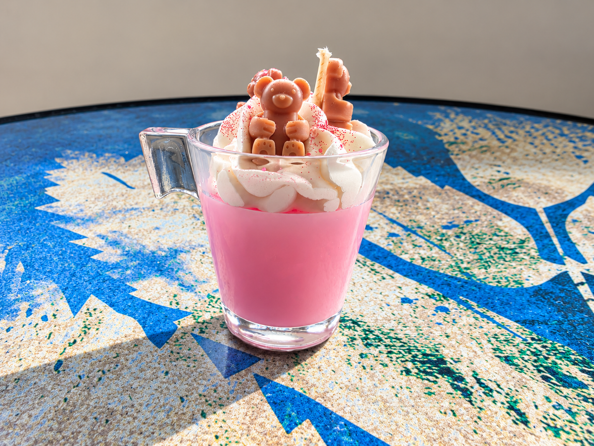 A pink beverage in a clear glass mug topped with whipped cream and small brown gummy bear candies, set on a blue and beige patterned table.