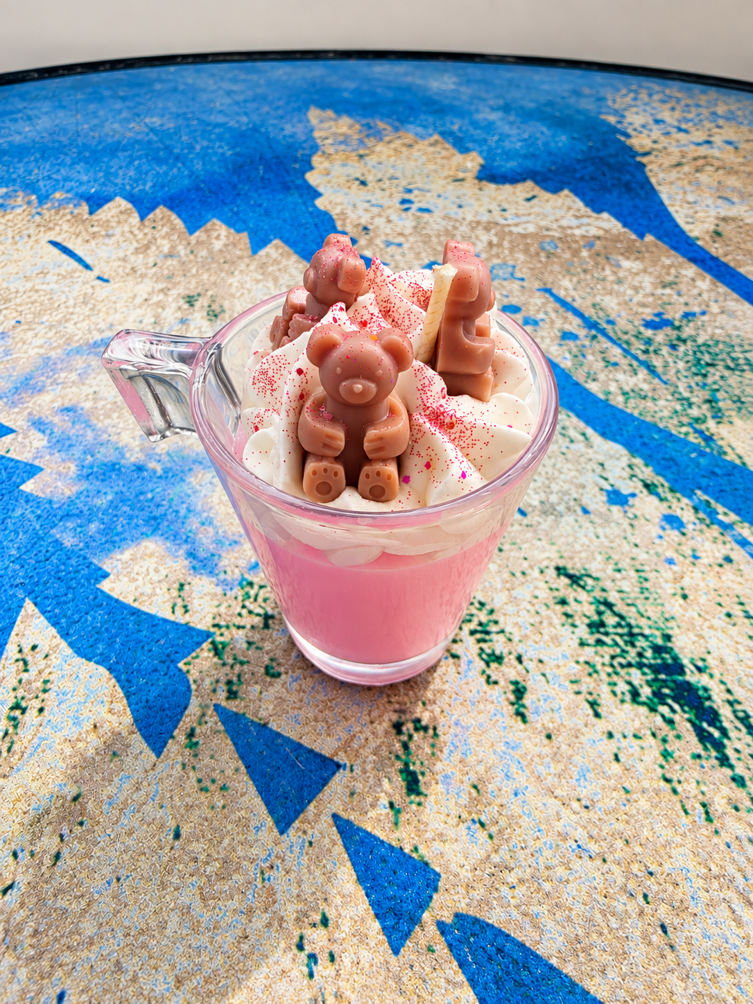 A pink dessert in a clear glass mug with whipped cream and chocolate decorations, placed on a blue and beige patterned surface.