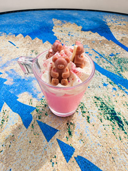 A pink dessert in a clear glass mug with whipped cream and chocolate decorations, placed on a blue and beige patterned surface.