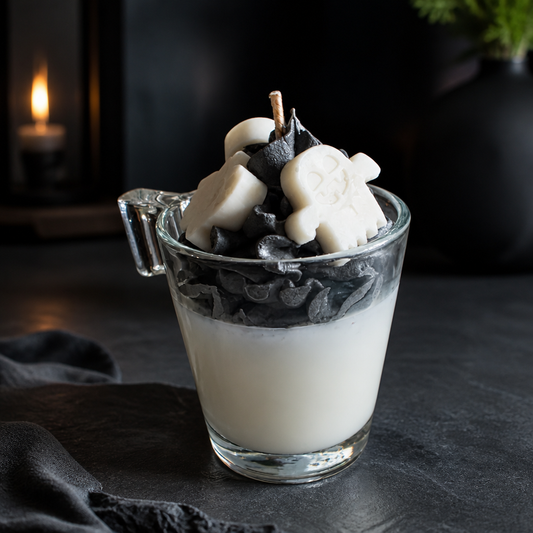 A layered candle in a clear glass mug with a handle, featuring a white base, dark charcoal black layers, and white skull-shaped wax decorations on top, set on a dark tabletop with a blurred candle in the background.