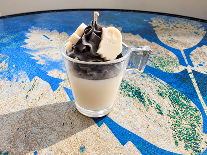 A novelty candle in a clear glass mug with a handle, filled with a light cream-colored layer and topped with dark brown whipped wax, chocolate swirls, and a white wax skull decoration, set on a blue and beige patterned table.