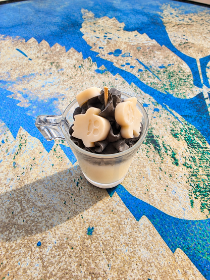 A small clear glass cup filled with layered cream and dark chocolate, topped with three pale beige ghost-shaped candies and a pretzel stick, set on a colorful blue and tan patterned surface.