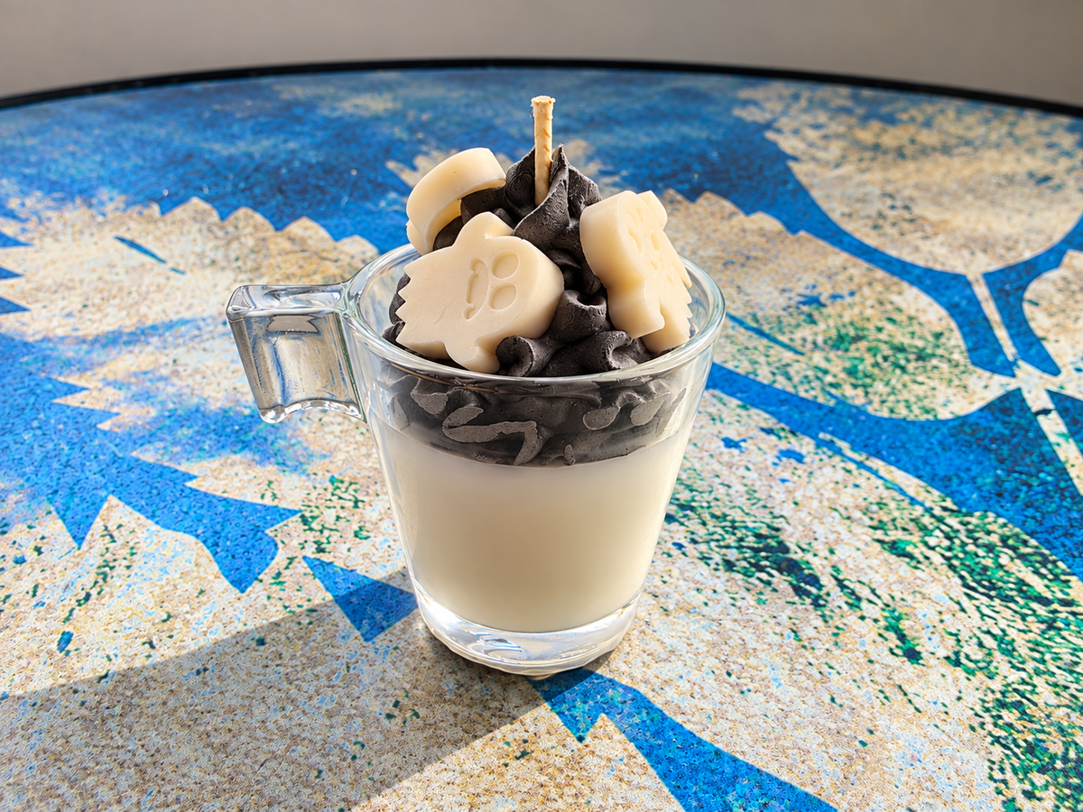 A layered dessert in a clear glass mug with a handle, featuring a creamy off-white base, dark chocolate chips, and pale cream ghost-shaped and banana-shaped toppings, set on a blue and beige patterned surface.