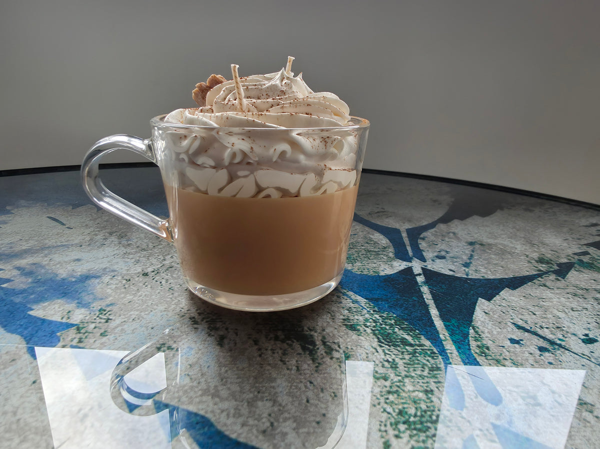 A clear glass mug filled with a light brown beverage, topped with whipped cream and chocolate shavings, sitting on a reflective surface with a gray background.