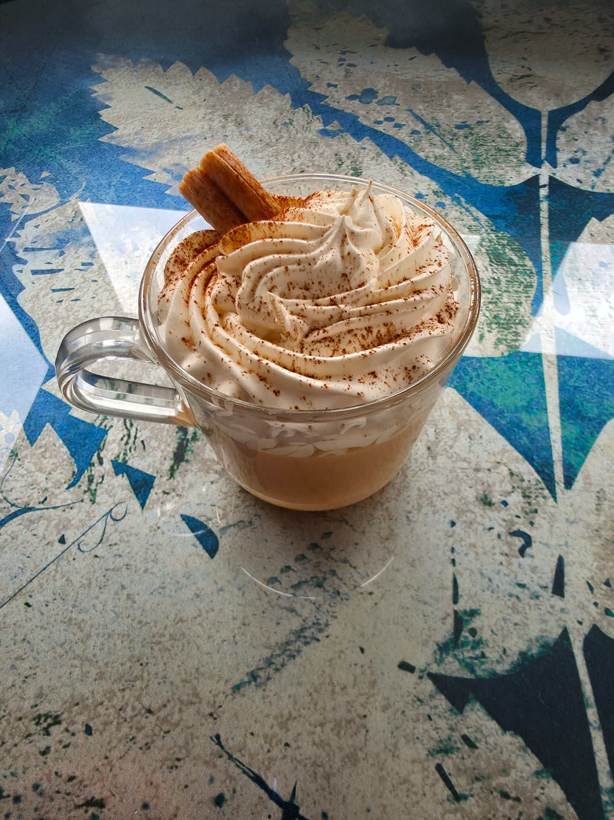 A clear glass mug filled with a hot cocoa or coffee drink topped with whipped cream and a cinnamon stick, set on a textured blue and gray surface.